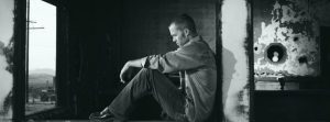 B&W image of man sitting, looking down & pondering.