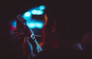 Person lighting a cigarette in her mouth at night with city lights in the back.