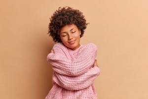 Woman hugging herself smiling in front of a orange background.