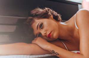 woman with makeup and tattoo leans on piano keys looking sad.