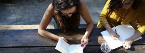 Two women sitting on a picnic bench one reading, one writing.