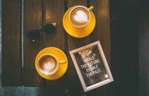 Two cappuccinos on yellow places, sunglasses and a framed sign on wood table.