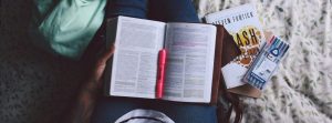 Close up of annotated book on persons lap sitting on a bed with a pink highlighter.