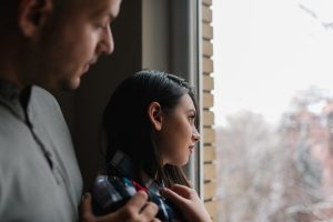 A woman looks out of a window on a cloudy day with a man in a gray shirt and his hand on her shoulder, comforting her.