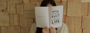 Woman holding a open book in front of her face that reads "Instead of choosing to merely exist, I urge you to experience life I urge you to live."