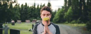 Man with praying hands holding a flower to hit face standing in a graveyard.
