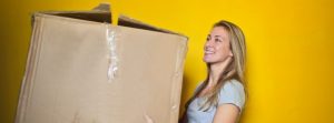Blonde woman holding a big cardboard box in front of yellow wall.