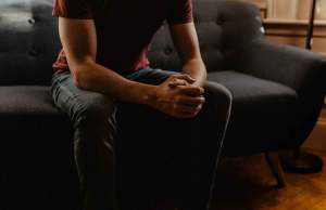 man in red t shirt and dark jeans sitting on grey couch holding his hands together.