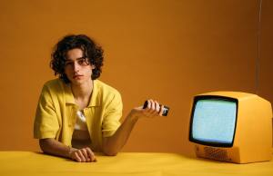 Bored teenage boy points remote control at yellow TV set.