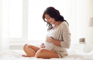 Pregnant woman smiling & holding her belly sitting on bed.