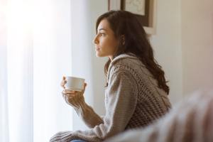 Woman looking forlorn in a sweater on the couch holding a cup.