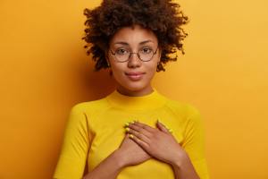 Woman in a yellow shirt holding her hands over her heart in front of a yellow/orange background.