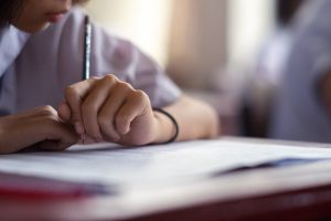 A student writes with a pencil on a piece of paper at a desk.
