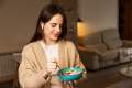 Woman in a tan sweater holding a blue ceramic bowl and eating granola with bluetooth earbuds in
