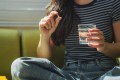 Young woman sitting comfortably on a sofa at home, holding a pill and a glass of water, preparing to take her medication