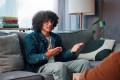 Young man sitting on couch at therapy session, arms out looking annoyed while sitting across from his therapist