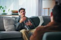 Young man on therapy couch, talking with his hands and looking content and engaged while speaking with male therapist, who we only see the back of, sitting across from him in a chair