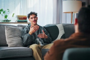 Young man on therapy couch, talking with his hands and looking content and engaged while speaking with male therapist, who we only see the back of, sitting across from him in a chair
