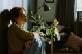A young woman with curly red hair sits on the floor hugging her knees to her chest, gazing downward with a distant expression in a dimly lit living room.