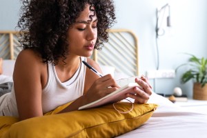 Woman lying on her bed writing in a journal, resting on a yellow pillow